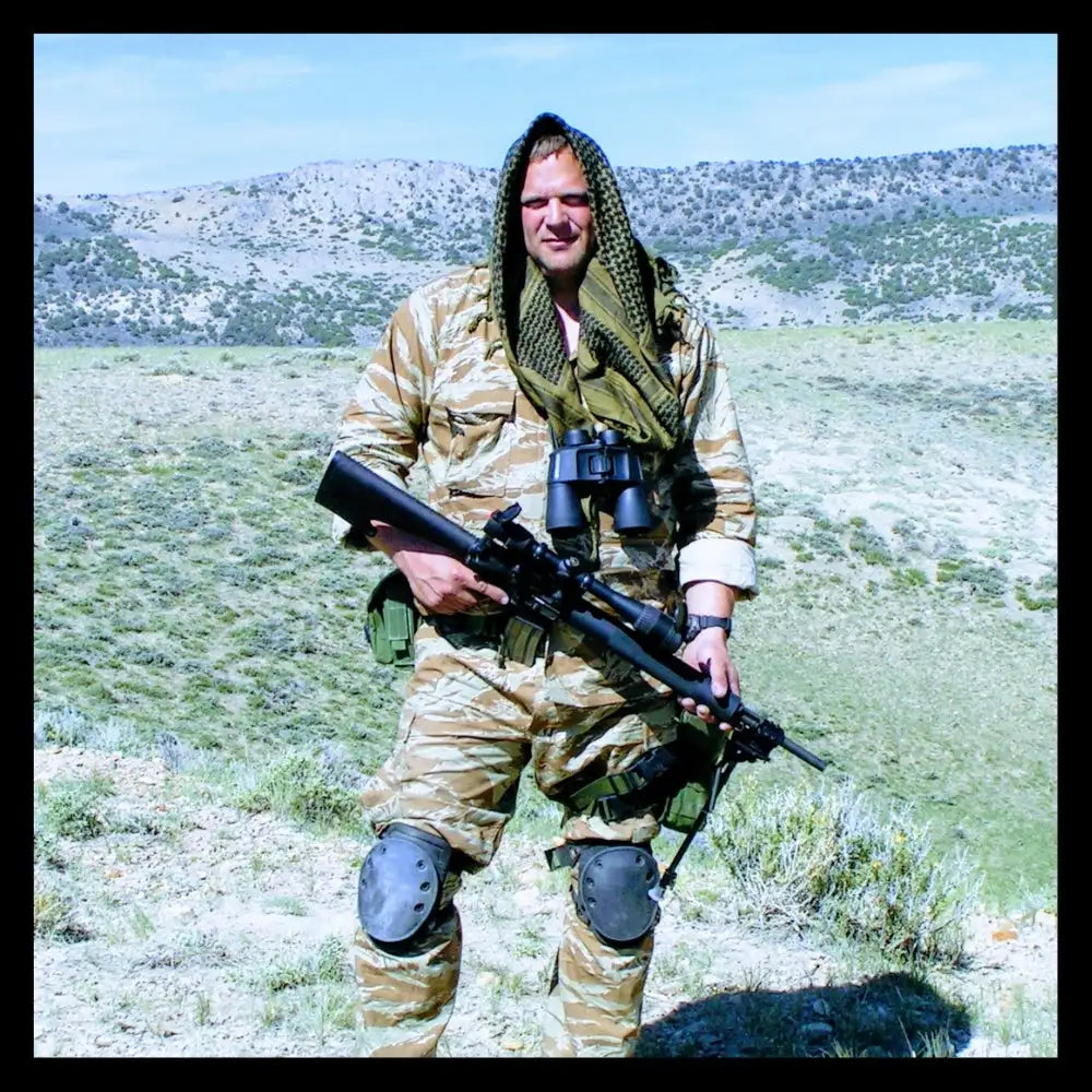Military camouflage uniform with black and tan pattern, knee pads, binoculars, rifle.